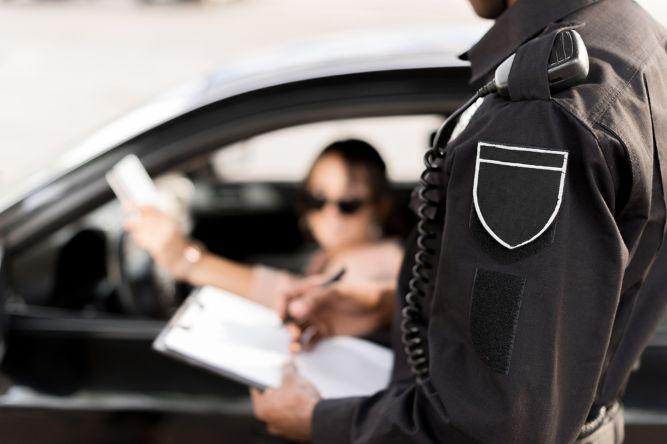 Cropped shot of policeman writing in clipboard and talking to woman sitting in car 